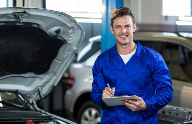garage-workers-peoplecreations Garage worker holding pen and clipboard beside car illustrates blog "Who Needs Garage Keepers Insurance? "