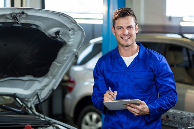 Garage worker holding pen and clipboard beside car illustrates blog "Who Needs Garage Keepers Insurance? "