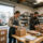 Realistic professional photograph of a small business owner and employee preparing holiday packages for delivery inside a modern California retail warehouse, cardboard shipping boxes, festive gift packages, delivery van visible through an open warehouse door, organized workspace, natural daylight, authentic commercial business environment, professional corporate photography, horizontal composition.